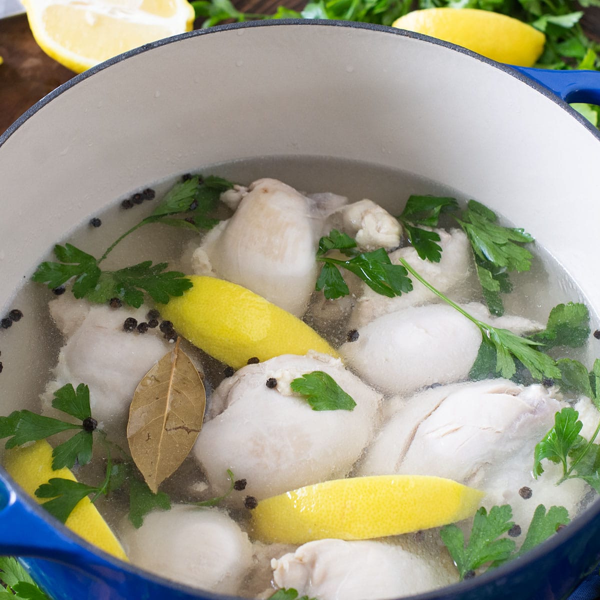Chicken thighs in a pot with stock, herbs, and lemon wedges.