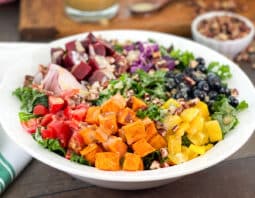 Salad in a white serving bowl with sweet potato, tomatoes, blueberries and more over kale.