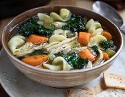 Bowl of chicken noodle soup with carrots and kale on a plate with crackers.
