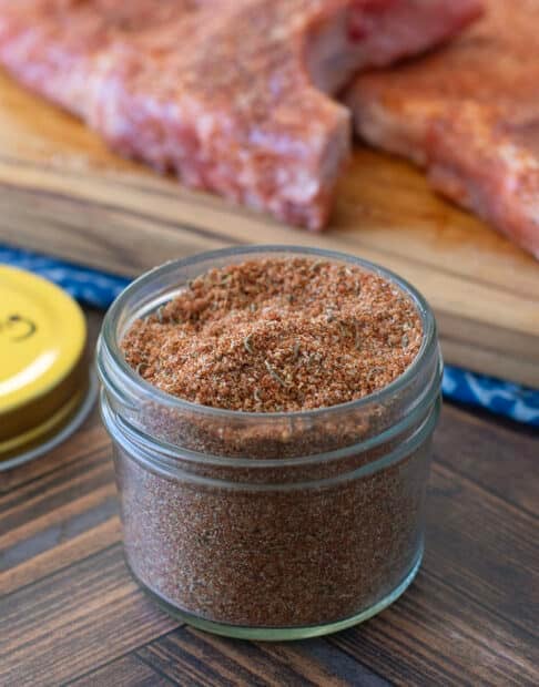 Small glass jar of pork chop seasoning blend with raw pork chops in background.