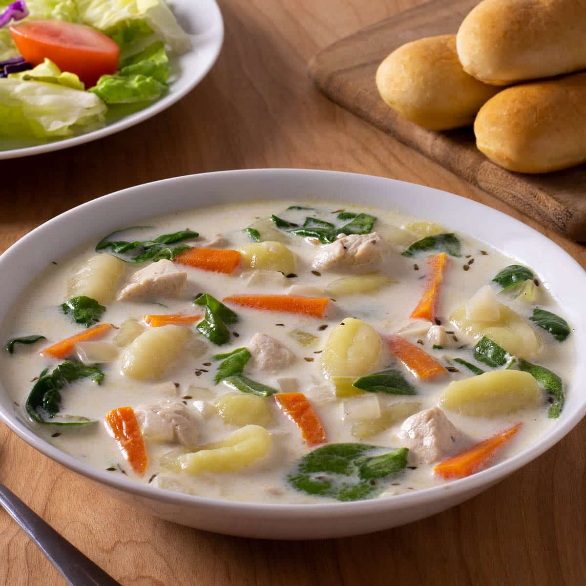 Bowl of chicken gnocchi soup with salad and breadsticks in background.