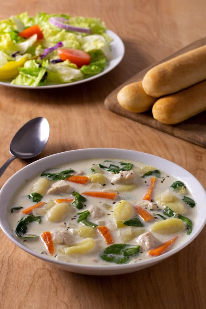 Bowl of chicken gnocchi soup with salad and breadsticks in background.