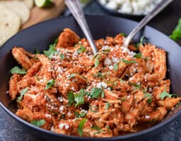 Black serving bowl of shredded chicken tinga, with tortillas and taco fixings in background.
