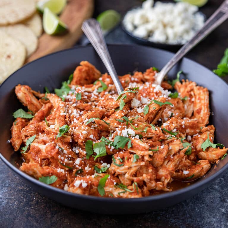 Black serving bowl of shredded chicken tinga, with tortillas and taco fixings in background.