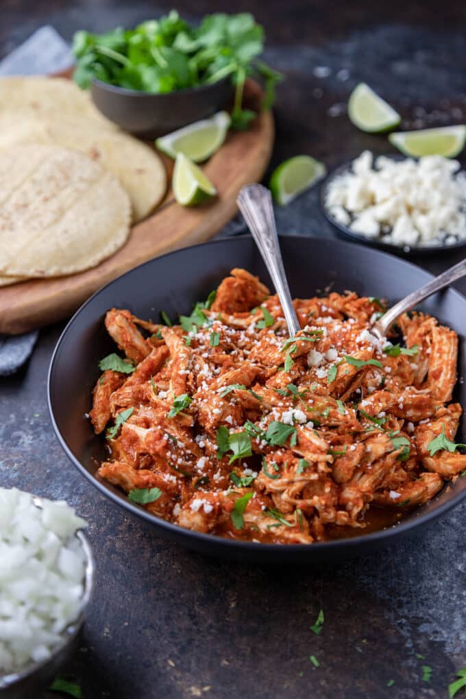 Black serving bowl of shredded chicken tinga, with tortillas and taco fixings in background.