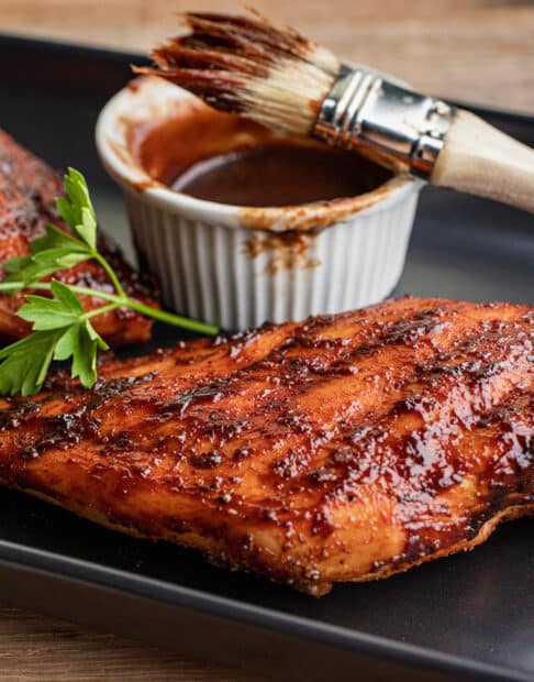 BBQ chicken breasts on a black platter with BBQ sauce dish and basting brush.