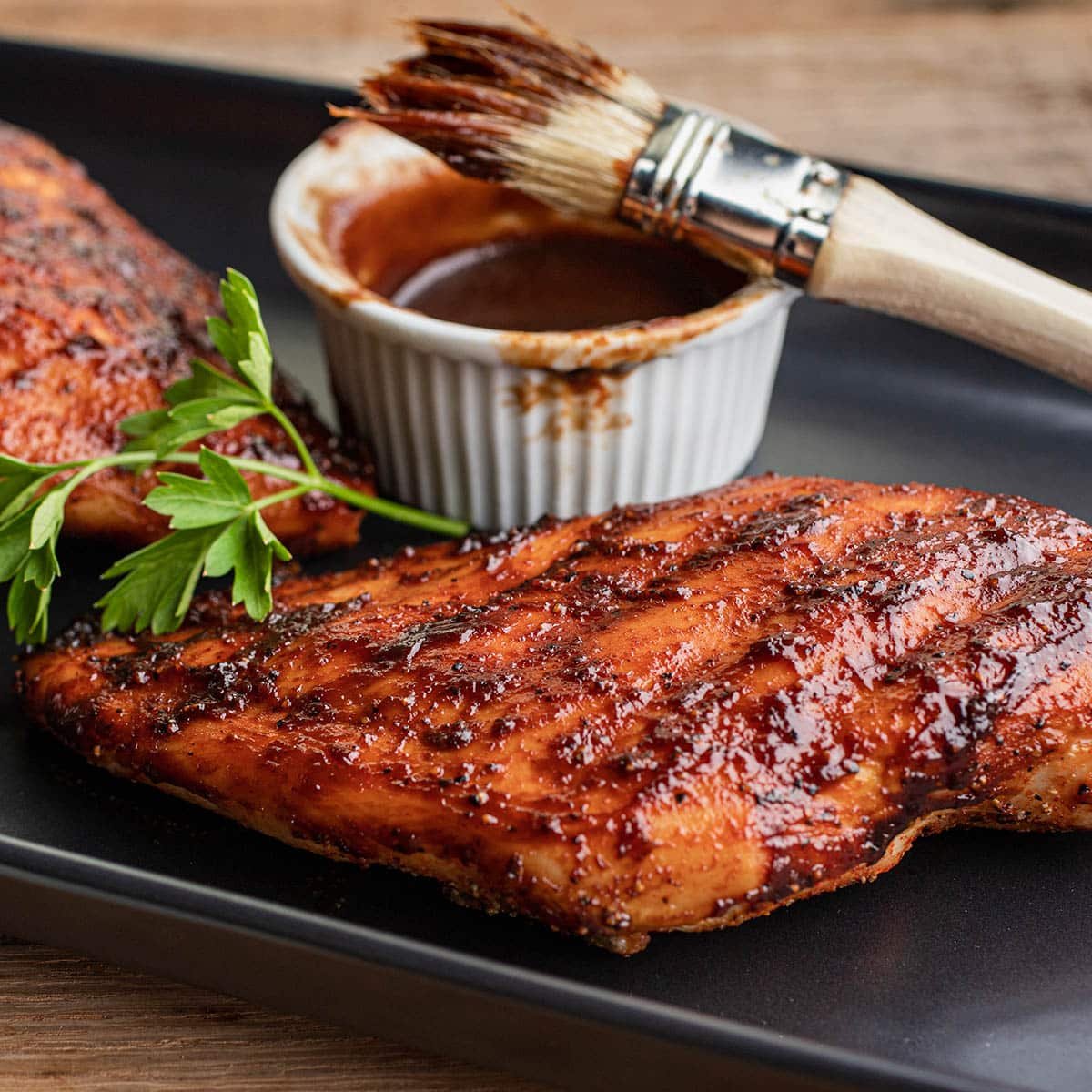 BBQ chicken breasts on a black platter with BBQ sauce dish and basting brush.