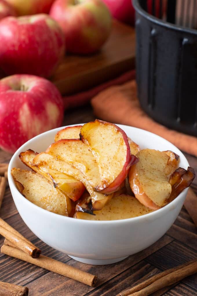 White bowl with sliced cooked apples. Whole apples and cinnamon sticks around.