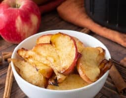 White bowl with sliced cooked apples. Whole apples and cinnamon sticks around.