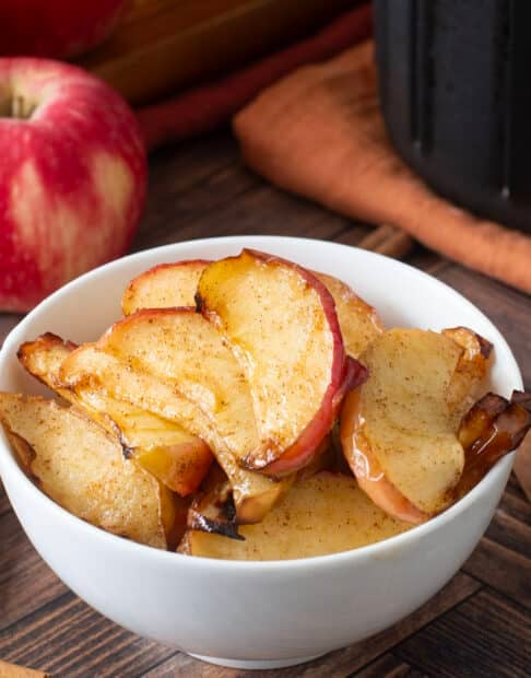 White bowl with sliced cooked apples. Whole apples and cinnamon sticks around.