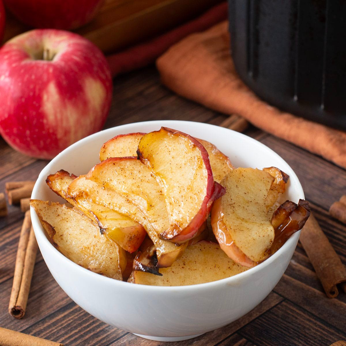 White bowl with sliced cooked apples. Whole apples and cinnamon sticks around.