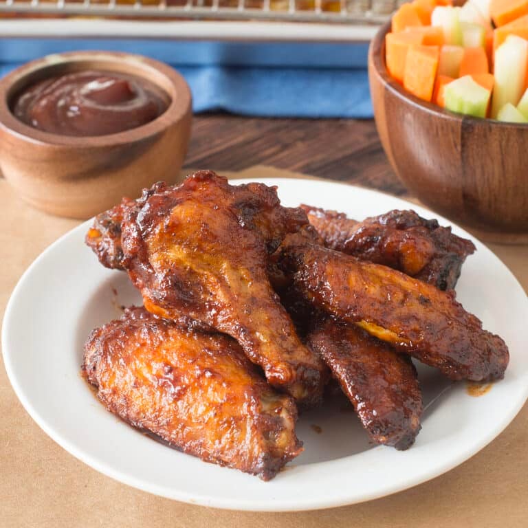 BBQ chicken wings on a white plate with BBQ sauce and veggie sticks behind.