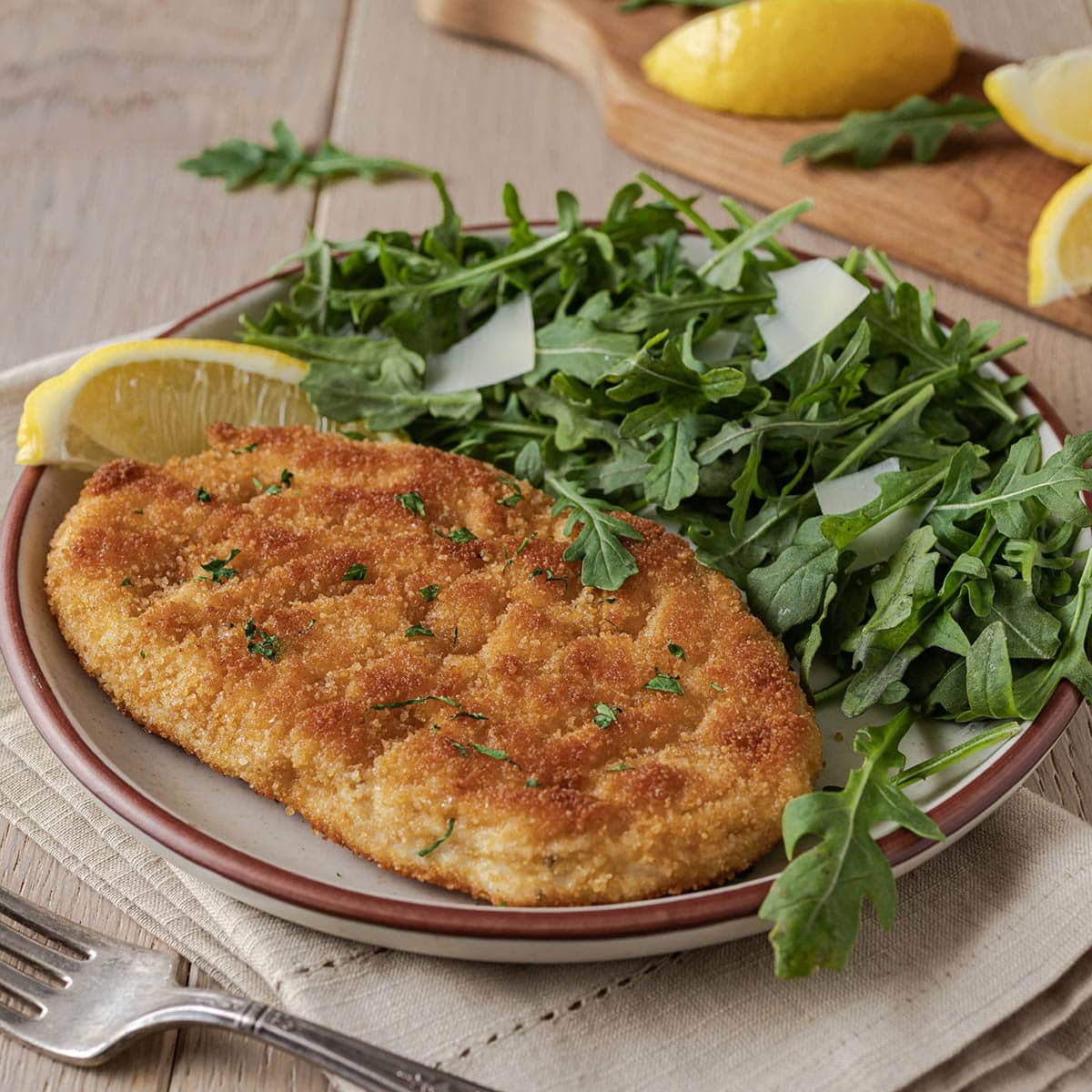 Breaded chicken cutlet on a plate with arugula salad and lemon wedge.