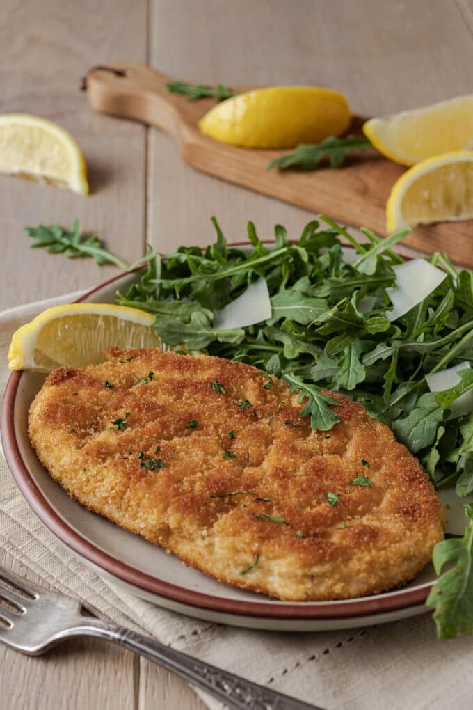 Breaded chicken cutlet on a plate with arugula salad and lemon wedge.