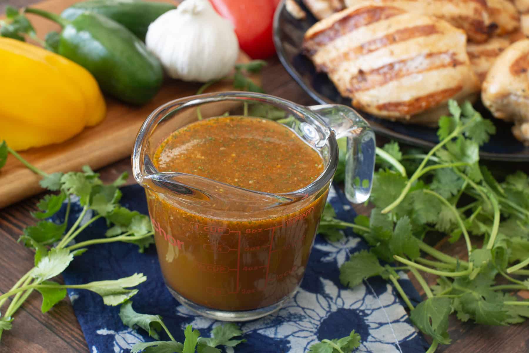 Glass measuring cup of Mexican chicken marinade surrounded by fresh ingredients like bell peppers.